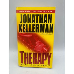 May include: A paperback book titled "THERAPY" by Jonathan Kellerman, a New York Times Bestseller. The cover is yellow with red accents and features the author's name in large, bold letters. The book is an Alex Delaware Novel.