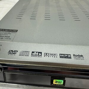 Pode incluir: Leitor de DVD prateado com painel frontal preto. Apresenta logotipos para DVD Video, Compact Disc Digital Audio, DTS Digital Out, Dolby Digital, MP3 MPEG Audio e Kodak Picture CD Compatible. Inclui um logotipo DVD-RWritable.