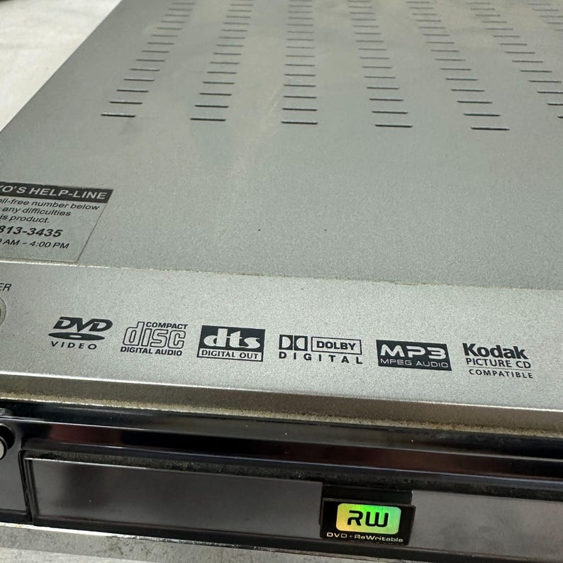 Pode incluir: Leitor de DVD prateado com painel frontal preto. Apresenta logotipos para DVD Video, Compact Disc Digital Audio, DTS Digital Out, Dolby Digital, MP3 MPEG Audio e Kodak Picture CD Compatible. Inclui um logotipo DVD-RWritable.