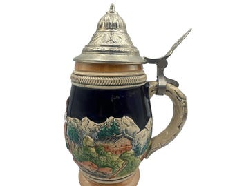 May include: A ceramic beer stein with a detailed mountain scene, featuring a house, trees, and figures. The stein has a dark blue body, a silver lid, and a decorative handle. The base and top are tan.