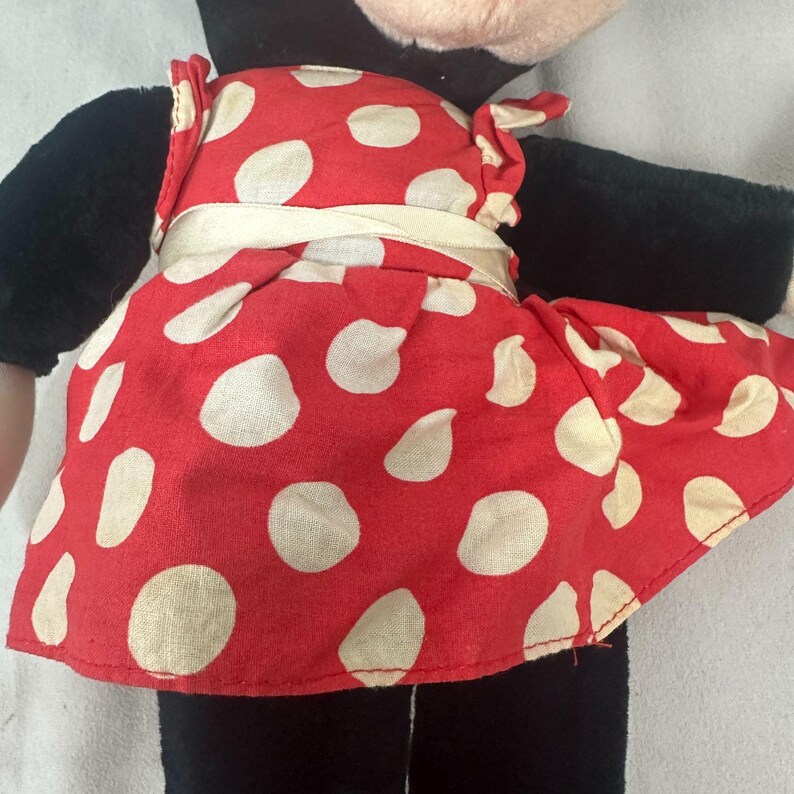 May include: A plush doll with a red dress adorned with white polka dots. The dress is cinched with a cream-coloured sash and worn over black arms and legs. The doll's face is partially visible, with a black head and ears.
