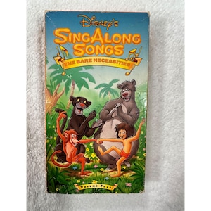 May include: A vintage VHS tape of Disney's Sing Along Songs: The Bare Necessities. The cover features cartoon characters Baloo the bear, Bagheera the panther, and Mowgli, with the text "Volume Four". The background shows a jungle scene.