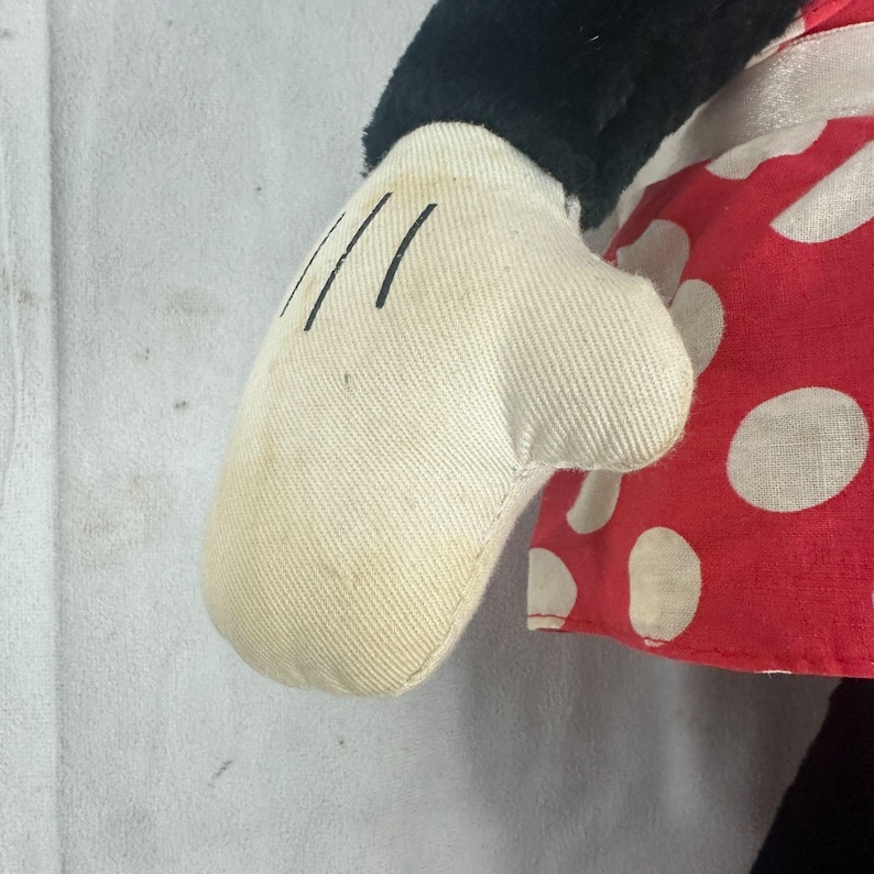 May include: Close-up of a plush toy's white glove with three black lines on the palm. The glove is slightly stained. The toy has a red and white polka dot dress and black sleeve.