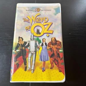 May include: A VHS tape of "The Wizard of Oz" featuring the film's iconic characters: Dorothy in a blue dress, the Tin Man, the Scarecrow, and the Cowardly Lion. The title is in yellow with a small illustration of the Emerald City.