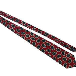 Peut inclure: Une cravate présentant un fond rouge profond avec un motif répétitif de formes cachemire bleu marine foncé soulignées de crème. La cravate est disposée en diagonale, montrant le pan large et l'extrémité étroite avec une étiquette visible.