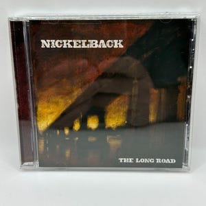 Op de afbeelding: Een Nickelback muziek-cd getiteld The Long Road in een doorzichtig plastic doosje. De hoes heeft een donkere, getextureerde achtergrond met tinten diep rood, oranje en zwart, met de silhouet van een brug boven het water.