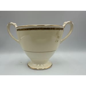 May include: Cream-colored ceramic bowl with two handles and a pedestal base. The bowl features gold trim along the rim, base, and a decorative band. The handles are curved and elegant, adding to the bowl's classic design.