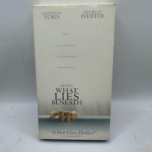 May include: A VHS tape box for the movie "What Lies Beneath" featuring Harrison Ford and Michelle Pfeiffer. The box has a white background with the movie title and cast names. The tagline reads "A First Class Thriller!"