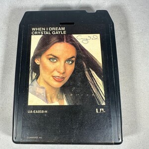 Puede incluir: Cinta de 8 pistas negra con el álbum "When I Dream" de Crystal Gayle. La portada muestra un retrato de Crystal Gayle con su firma. La cinta tiene la etiqueta "UA-EA858-H" y el logotipo de UA.
