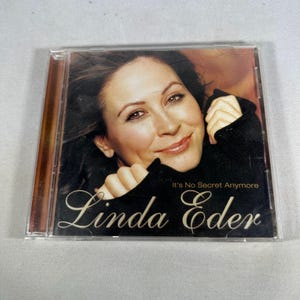 May include: A compact disc case featuring a woman's portrait with the text "It's No Secret Anymore" and "Linda Eder" in a stylized font. The background has a warm, brown tone.