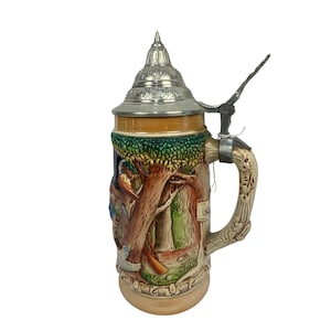 May include: A ceramic beer stein featuring a relief-moulded forest scene with trees and foliage in shades of green and brown. The vessel has a metallic lid with a pointed finial and a textured handle shaped like a tree branch, standing upright.