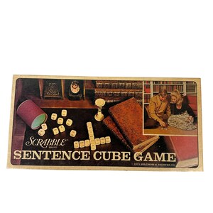 Pode incluir: Uma caixa do jogo Scrabble Sentence Cube Game de 1971 com um gráfico de dados, um copo roxo e dois livros encadernados em couro. A tampa exibe o texto Scrabble Brand Sentence Cube Game e uma foto de duas pessoas jogando o jogo.