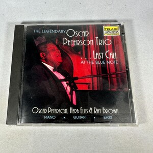 May include: A CD case for "The Legendary Oscar Peterson Trio - Last Call at the Blue Note." The cover features a portrait of Oscar Peterson against a red backdrop. The musicians' names and instruments are printed below the image.