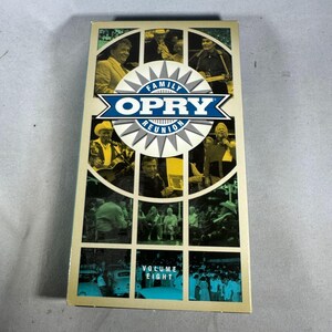 Pode incluir: Uma fita VHS vintage de "Family Opry Reunion" Volume Eight. A capa apresenta uma colagem de artistas em um padrão de grade, com o logotipo Opry em azul e branco. A fita tem um fundo creme.