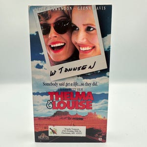 Puede incluir: Una cinta VHS de la película "Thelma & Louise" con Susan Sarandon y Geena Davis. La portada muestra una foto de las actrices y el título de la película. El fondo muestra un paisaje desértico con el texto "Somebody said get a life...so they did."