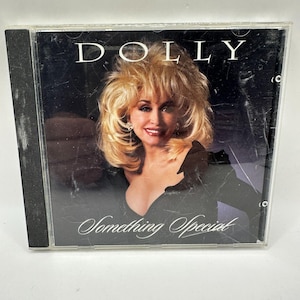 Puede incluir: Una caja de CD con Dolly Parton. La portada tiene un fondo negro con la palabra "DOLLY" en la parte superior. La imagen muestra a Dolly Parton con cabello rubio y una blusa negra. El título "Something Special" está escrito en cursiva.