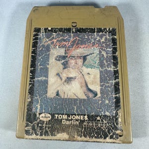 May include: An 8-track tape featuring Tom Jones. The album cover shows a portrait of Tom Jones wearing a hat. The tape is housed in a tan plastic cartridge with the album title "Darlin'" and the PolyGram Records logo.