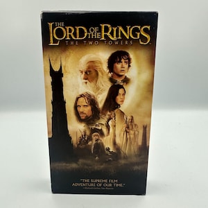 Può includere: Una videocassetta VHS del film "Il Signore degli Anelli: Le Due Torri". La copertina presenta un collage di personaggi e una silhouette di torre scura su uno sfondo color seppia. Il testo "THE LORD OF THE RINGS THE TWO TOWERS" è in alto.