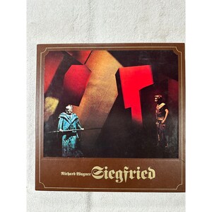 May include: A brown book cover featuring a stage production image. The image shows two costumed figures on a stage with large red and brown geometric shapes. The text "Richard Wagner Siegfried" is printed in gold.