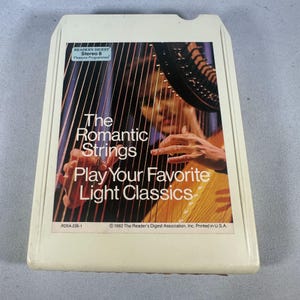 May include: A vintage Reader's Digest Stereo 8 tape titled "The Romantic Strings Play Your Favorite Light Classics." The tape features a graphic of a person playing a harp. The tape is off-white with black and orange text.