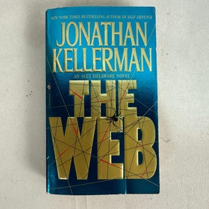 May include: A hardcover book titled "The Web" by Jonathan Kellerman. The cover is a gradient of blue and gold, with a spider graphic. Text includes "New York Times Bestselling Author" and "An Alex Delaware Novel."