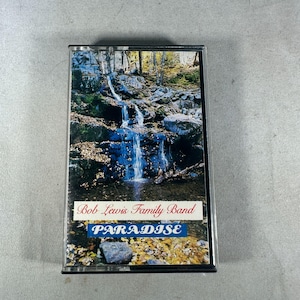 Puede incluir: Un casete vintage con el álbum "Paradise" de Bob Lewis Family Band. La portada muestra una escena de cascada con el nombre de la banda y el título del álbum en texto blanco y azul. Un formato musical nostálgico.
