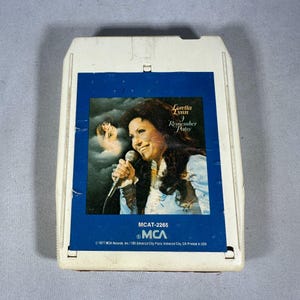 May include: An 8-track tape featuring Loretta Lynn's album "Remember Patsy." The tape has a white plastic casing with a blue label. The label features a portrait of Loretta Lynn holding a microphone, with the album title and MCA Records branding.