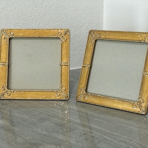 May include: Two square picture frames with ornate gold-colored borders. The frames have a light yellow finish with decorative scrollwork at the corners and along the sides. The frames are empty, with a light gray backing.
