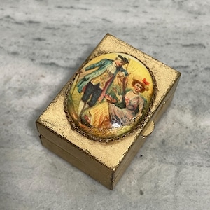 May include: A rectangular gold-toned metal box featuring an oval inset on the lid. The inset displays a colorful illustration of two figures in period clothing, with one standing and one seated, surrounded by a textured gold rope-style border.