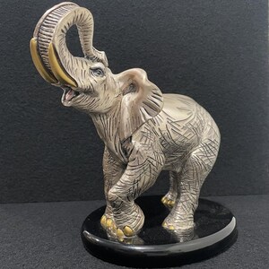 May include: A silver and gold elephant sculpture with its trunk raised in the air. The elephant is standing on a black base.