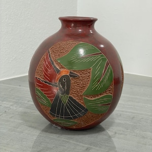 May include: A round, red vase with a detailed, carved design. The design features a bird with a black body, orange head covering, and a black skirt, surrounded by green leaves. The vase has a smooth, glossy finish.