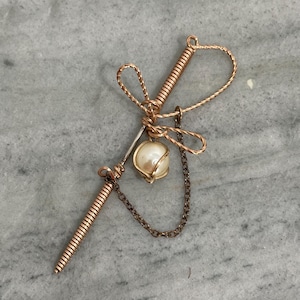 May include: A handcrafted copper-colored wire hair accessory. The design features a decorative bow, a pearl, and a chain. The accessory has a unique, artistic appearance, suitable for special occasions or everyday wear.