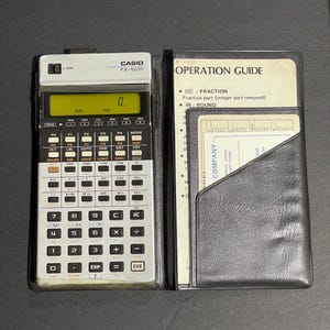 May include: A Casio FX-501P scientific calculator with a black and silver design, displayed open with its operation guide and a black leatherette case. The calculator's display shows a zero. The calculator has many buttons for various functions.