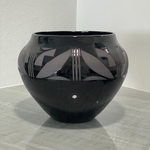 May include: A glossy black ceramic bowl with a wide, rounded body and a narrow opening. The exterior features a repeating pattern of matte grey, leaf-shaped motifs and vertical lines encircling the upper portion of the vessel.
