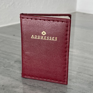 May include: A small, burgundy address book with gold lettering that reads "ADDRESSES". The book is rectangular and appears to be made of leather or a similar material. The cover has a stitched border.