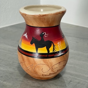 May include: A hand-painted ceramic vase with a Southwestern design. The vase features a horse and rider silhouette against a sunset backdrop of red, yellow, and orange. The vase has a light brown base with etched patterns and a cream-colored rim.