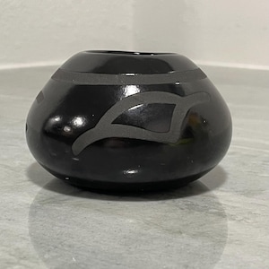 May include: A small, round, black ceramic vase with a glossy finish. The vase features a gray, geometric design around the top and a stylized shape on the side. The vase is a decorative item.