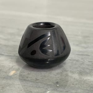 May include: A small, polished black ceramic vessel with a rounded base and a wide, circular opening at the top. The exterior surface features a series of etched, matte-finish geometric patterns that contrast with the glossy black finish of the pot.