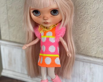 Può includere: Una bambola con lunghi capelli rosa chiaro e occhi blu. La bambola indossa un vestito rosa e arancione brillante con disegni a stella bianchi, un fiocco giallo con pois dorati e stivali rosa. L'abito ha un bordo giallo.