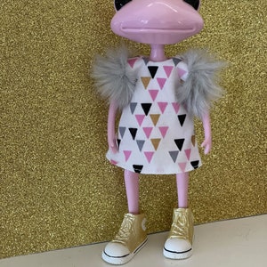 May include: A pink frog figurine wearing a white dress with pink, black, and gold triangle pattern. The frog has gray fur shoulder accents and gold high-top sneakers with white soles. The background is a gold glitter surface.