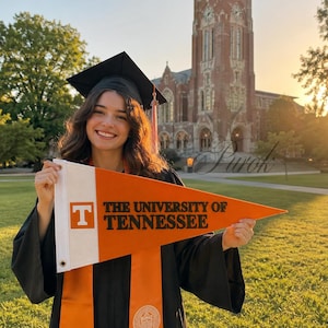 Puede incluir: Una persona con toga y birrete sostiene una bandera naranja y blanca con la inscripción "THE UNIVERSITY OF TENNESSEE" y una "T". El fondo muestra un edificio universitario, lo que sugiere una ceremonia de graduación.