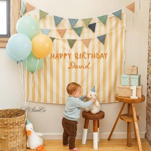 May include: A birthday scene featuring a yellow and white striped backdrop with the text "HAPPY BIRTHDAY David". A baby in a blue sweater and brown trousers interacts with a stuffed lamb wearing a party hat. Balloons and gifts are also present.
