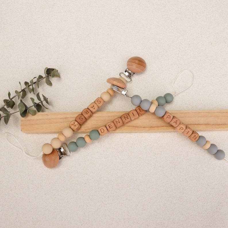 Puede incluir: Tres chupeteros personalizados para beb&eacute;s con cuentas de madera con los nombres "SARSA", "DENNIE" y "PAPIN". Los clips presentan una mezcla de madera natural, cuentas grises y turquesas, con un clip de madera y un lazo.