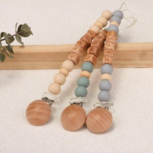 Puede incluir: Tres clips de chupete de madera con cuentas de madera y clips de metal. Los clips tienen nombres escritos con cuentas de letras de madera. Las cuentas son en tonos beige, gris y verde azulado.