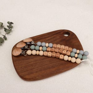 Puede incluir: Dos clips para chupete de beb&eacute; con cuentas de madera y silicona en colores neutros. Los clips tienen discos de madera y cierres met&aacute;licos. Un clip deletrea "PAPPIN DENNIE SARS". Los clips est&aacute;n sobre una tabla de cortar de madera.