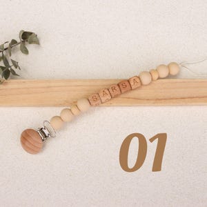 Puede incluir: Pinza para chupete de beb&eacute; de madera con el nombre "SARSA" escrito con cuentas de letras de madera. El clip tiene un disco de madera y un cierre met&aacute;lico. El n&uacute;mero "01" est&aacute; en la esquina inferior derecha.