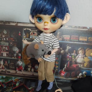 Puede incluir: Una muñeca con cabello azul y ojos azules grandes, vestida con una camisa a rayas y pantalones color caqui, sosteniendo una rata de peluche gris. La muñeca tiene una curita en la nariz. El fondo presenta una escena de una tienda de juguetes vintage.