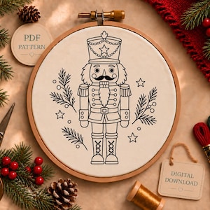 Könnte beinhalten: Ein runder hölzerner Stickrahmen mit cremefarbenem Stoff, auf dem ein schwarzes Linienmuster eines Nussknacker-Soldaten, umgeben von Tannenzweigen und Sternen, abgebildet ist. Der Text PDF PATTERN und DIGITAL DOWNLOAD ist sichtbar.
