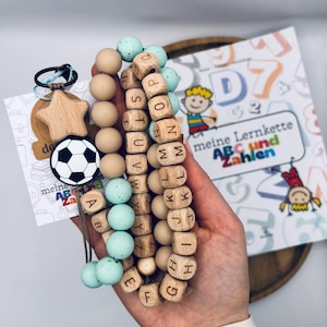 Könnte beinhalten: Eine Lernkette mit hölzernen Buchstabenperlen, glatten beigen Silikonperlen und gesprenkelten mintgrünen Silikonperlen. Ein hölzerner Sternanhänger und ein schwarz-weißer Fußballanhänger sind an einem Schlüsselring mit einer Karte meine Lernkette befestigt.