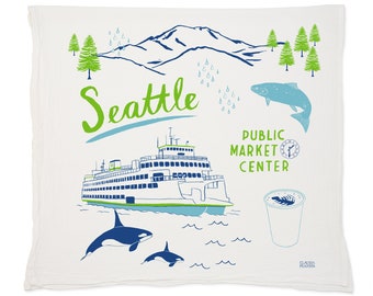 May include: A white tea towel with a blue and green illustration of Seattle landmarks. The illustration includes a ferry, a salmon, a coffee cup, orcas, and the text "Seattle", "Public Market Centre", and "Claudia Pearson".